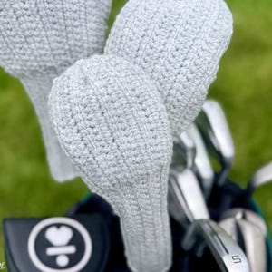 Pode incluir: Close-up de um saco de golfe com vários tacos e três capas de crochê cinza claro. As capas são feitas de fio texturizado e cobrem as cabeças dos tacos. O fundo é grama verde desfocada.
