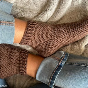 May include: Close-up of brown knitted socks with a textured pattern. The socks are worn with blue jeans. The socks have a ribbed cuff and cover the feet and ankles. The image is taken indoors.