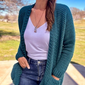 May include: A teal blue cardigan with pockets and a white vest top underneath. The cardigan is made of a soft, textured yarn and has a relaxed fit. The model is wearing blue jeans.