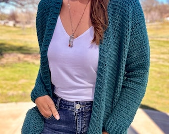 May include: A teal blue cardigan with pockets and a white tank top underneath. The cardigan is made of a soft, textured yarn and has a relaxed fit. The model is wearing blue jeans.