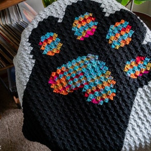May include: A handmade crochet blanket featuring a large paw print design. The paw print is made with colorful squares of orange, pink, and blue. The blanket has a black paw print on a gray background. The blanket is made of soft yarn.