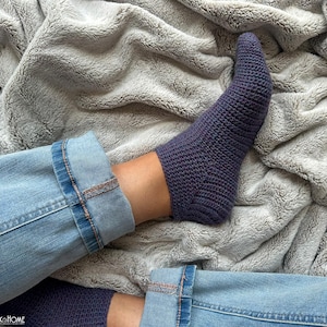 May include: A close-up shot of a person's legs, wearing blue jeans and a dark purple, knitted sock. The sock is textured and covers the foot and ankle. The person is resting on a soft, gray blanket.