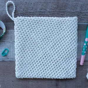 May include: A white crocheted pot holder with a loop handle. The pot holder is made with a textured stitch pattern. The image also shows a crochet hook, yarn, scissors, and a stitch marker.