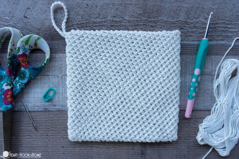 May include: A white crocheted pot holder with a loop handle. The pot holder is made with a textured stitch pattern. The image also shows a crochet hook, yarn, scissors, and a stitch marker.