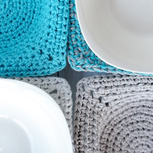 May include: Two crocheted square coasters in gray and turquoise blue, with white bowls on top. The coasters are made with a simple stitch pattern and have a slightly textured surface.