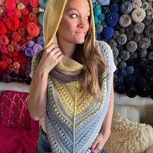 May include: A woman wearing a crocheted hooded shawl with a yellow, gray, and brown color scheme. The shawl is made with a textured stitch and has a triangular shape. The woman is standing in front of a wall of yarn balls.