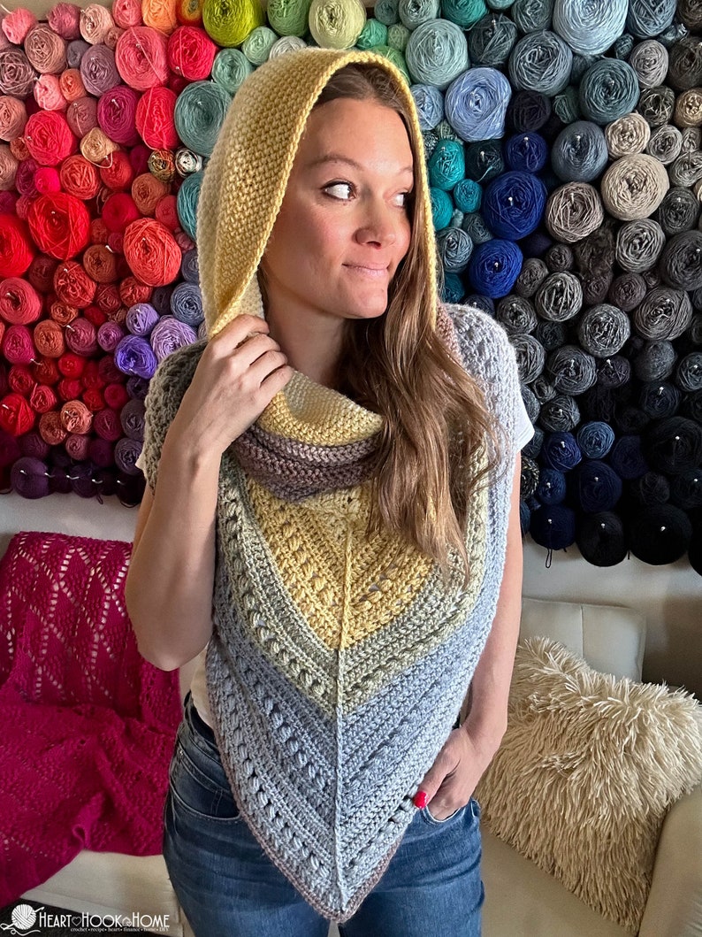 May include: A woman wearing a crocheted hooded shawl with a yellow, gray, and brown color scheme. The shawl is made with a textured stitch and has a triangular shape. The woman is standing in front of a wall of yarn balls.