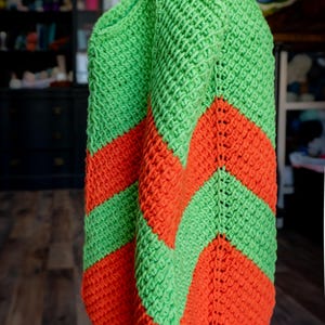 May include: A handmade crochet bag in bright green and orange. The bag features a chevron pattern in the two colors. The bag is made of a thick yarn, giving it a textured appearance. The bag is likely a tote or market bag.