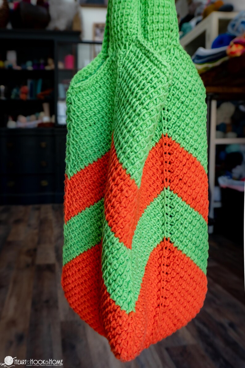 May include: A handmade crochet bag in bright green and orange. The bag features a chevron pattern in the two colors. The bag is made of a thick yarn, giving it a textured appearance. The bag is likely a tote or market bag.