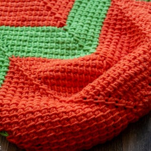 May include: Close-up of a handmade crochet blanket in vibrant orange and green. The blanket features a textured stitch pattern and a geometric design. The warm colors and cozy texture suggest a handmade home decor item.