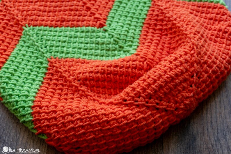 May include: Close-up of a handmade crochet blanket in vibrant orange and green. The blanket features a textured stitch pattern and a geometric design. The warm colors and cozy texture suggest a handmade home decor item.