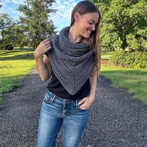 May include: A gray triangular shawl with a crocheted design. The shawl is worn by a person with long brown hair, a black shirt, and blue jeans.