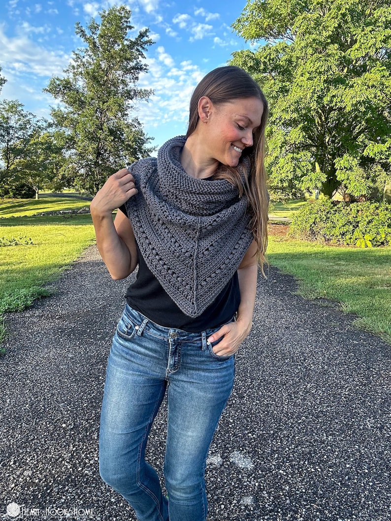 May include: A gray triangular shawl with a crocheted design. The shawl is worn by a person with long brown hair, a black shirt, and blue jeans.