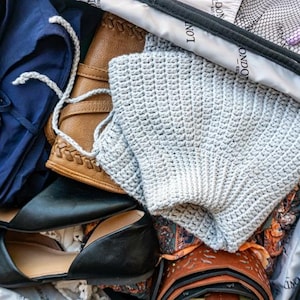 Puede incluir: Maleta abierta llena de art&iacute;culos de viaje. Incluye un su&eacute;ter de ganchillo gris claro, una blusa azul marino, bailarinas negras, una cartera de cuero marr&oacute;n y un secador de pelo. La maleta est&aacute; forrada con una tela estampada.