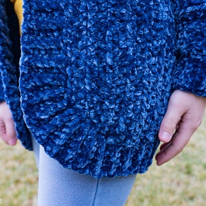 May include: A close-up of a blue fuzzy cardigan with a soft, textured fabric. The cardigan is open in the front and the sleeves are long.