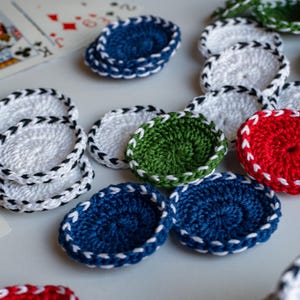 Op de afbeelding: Handgemaakte gehaakte pokerchips in wit, blauw, groen en rood. Elke ronde chip heeft een contrasterende rand. De chips zijn verspreid, sommige opgestapeld. Speelkaarten zijn zichtbaar op de achtergrond.