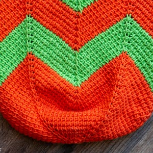 May include: Close-up of a crocheted item with a chevron pattern. The design features alternating bands of bright orange and green. The texture of the crochet stitches is clearly visible, creating a tactile appearance. The item is likely a bag or a similar container.
