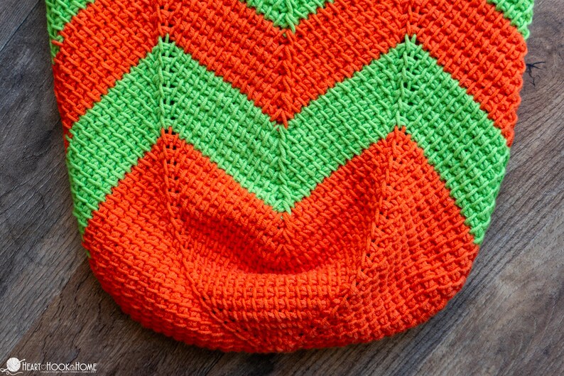 May include: Close-up of a crocheted item with a chevron pattern. The design features alternating bands of bright orange and green. The texture of the crochet stitches is clearly visible, creating a tactile appearance. The item is likely a bag or a similar container.