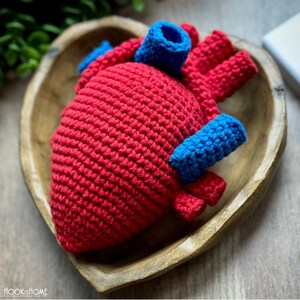 May include: A crocheted anatomical heart model featuring a textured red main body with prominent blue yarn vessels attached to the top and sides. The piece is crafted with visible yarn stitches and sits within a heart-shaped wooden bowl.
