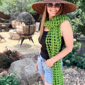 May include: A brown wide-brimmed straw hat, a bright green open-weave crochet scarf, a black sleeveless top, and light blue denim shorts. The scarf features a repeating geometric pattern with circular openings throughout the length of the fabric.