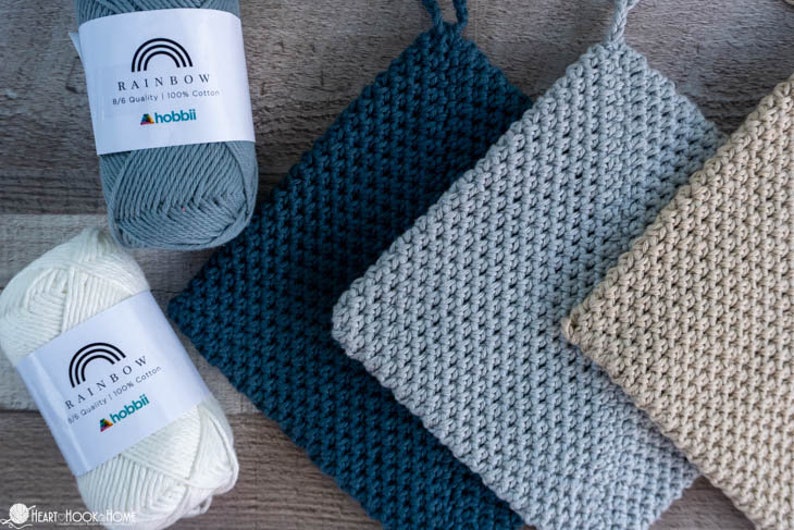 May include: Three crocheted pot holders in teal, grey, and beige. The pot holders are made with a textured stitch and are stacked on top of each other. Three balls of yarn in white, grey, and teal are also shown in the image. The yarn labels read "Rainbow 8/8 Quality 100% Cotton" and "Hobbii."