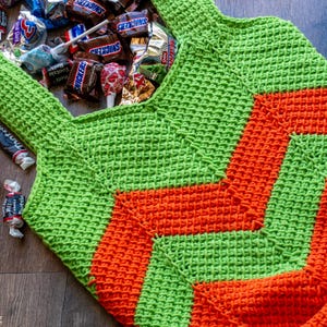 May include: A handmade green and orange crochet bag filled with various candies. The bag features a geometric design and is placed on a wooden surface. The bag is designed to carry candy.