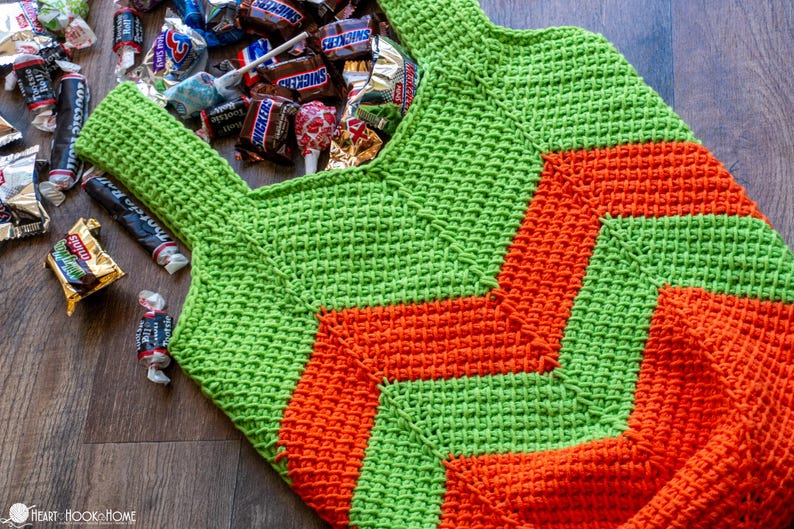 May include: A handmade green and orange crochet bag filled with various candies. The bag features a geometric design and is placed on a wooden surface. The bag is designed to carry candy.