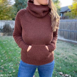May include: A brown long-sleeved sweater featuring a textured waffle-knit pattern, a high cowl neckline, and a large front kangaroo pocket. The garment is paired with blue denim jeans, showcasing a consistent knit construction throughout the piece.
