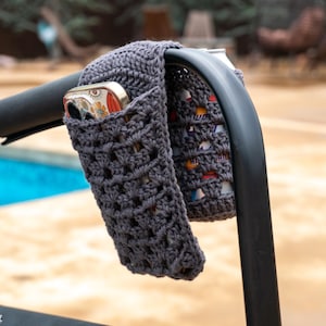 May include: Gray crocheted phone holder attached to a black chair arm. The holder is made with a simple stitch pattern and has a pocket for a phone.