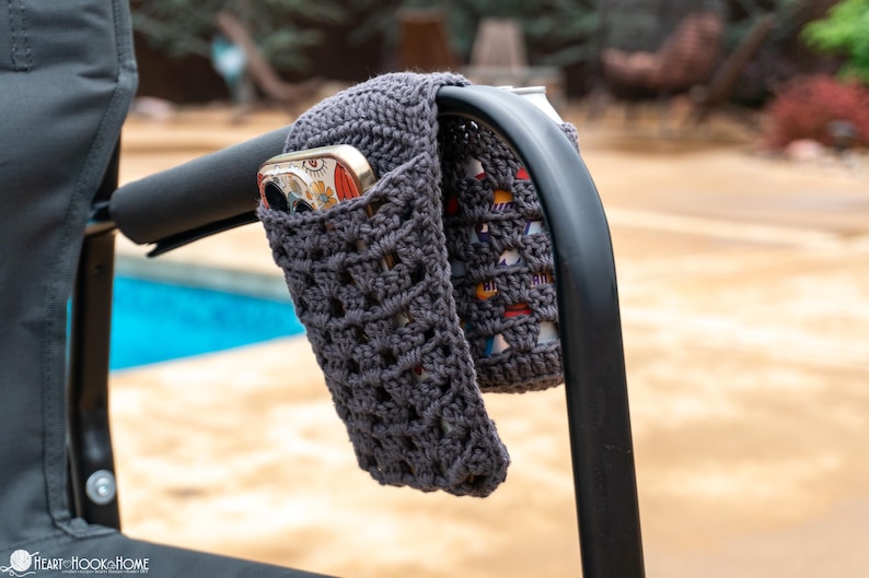 May include: Gray crocheted phone holder attached to a black chair arm. The holder is made with a simple stitch pattern and has a pocket for a phone.