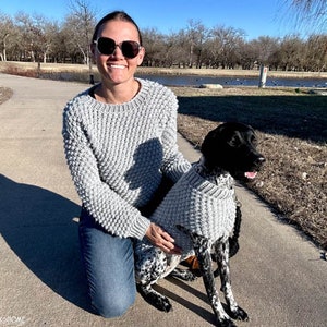 May include: A woman wearing a grey knitted sweater and blue jeans is kneeling next to a black and white dog wearing a matching grey knitted sweater. The dog is looking to the right of the frame. The woman is wearing sunglasses and smiling.