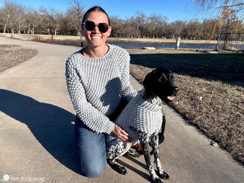 May include: A woman wearing a grey knitted sweater and blue jeans is kneeling next to a black and white dog wearing a matching grey knitted sweater. The dog is looking to the right of the frame. The woman is wearing sunglasses and smiling.