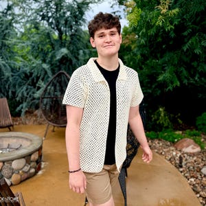 May include: A cream-coloured, short-sleeved, open-front crochet shirt featuring a classic collar and a repeating diamond-shaped mesh pattern. The garment is worn over a plain black t-shirt and paired with tan shorts, showcasing a textured, lightweight design.