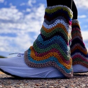 May include: A pair of white athletic sneakers worn with multicolored crochet boot cuffs featuring a chevron pattern in shades of blue, green, yellow, brown, and white. The cuffs extend above the ankle, and the text Heart Hook Home is visible.