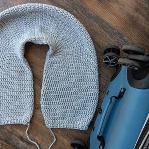 Puede incluir: Una almohada de viaje de ganchillo gris claro en forma de U, sobre una superficie de madera junto a una maleta azul. La almohada est&aacute; hecha a mano y parece suave. La maleta tiene ruedas negras y un asa.