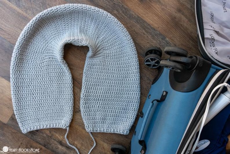 Puede incluir: Una almohada de viaje de ganchillo gris claro en forma de U, sobre una superficie de madera junto a una maleta azul. La almohada est&aacute; hecha a mano y parece suave. La maleta tiene ruedas negras y un asa.