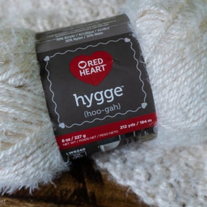 May include: A skein of white yarn with a fuzzy texture. The yarn label reads "Red Heart Hygge (hoo-gah)" and indicates that the yarn is 70% acrylic, 20% acrylic, and 10% nylon. The label also states that the skein contains 8 ounces (227 grams) of yarn and 212 yards (194 meters).