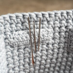 May include: Close-up of a light gray crocheted basket with three silver needles inserted. The basket has a textured, woven appearance, and the needles are positioned vertically. The image highlights the texture and craftsmanship of the handmade item.