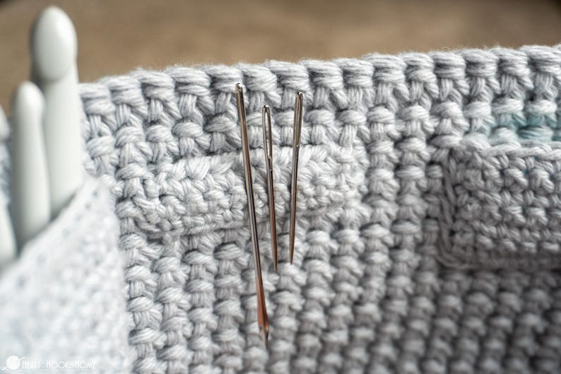 May include: Close-up of a light gray crocheted basket with three silver needles inserted. The basket has a textured, woven appearance, and the needles are positioned vertically. The image highlights the texture and craftsmanship of the handmade item.