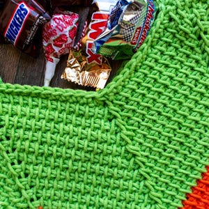 May include: Close-up of a bright green crocheted item with an orange accent. Candy bars and a lollipop are nestled in the opening. The crochet work has a textured, woven appearance, suggesting a handmade craft project.