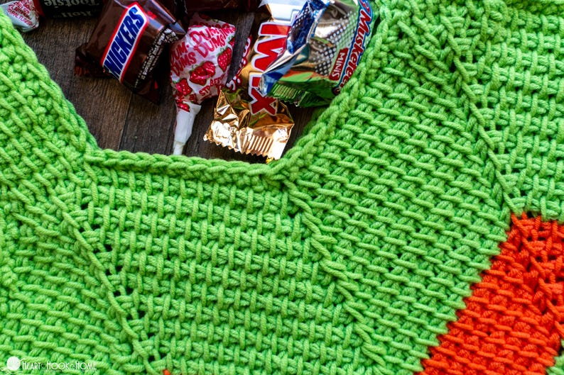 May include: Close-up of a bright green crocheted item with an orange accent. Candy bars and a lollipop are nestled in the opening. The crochet work has a textured, woven appearance, suggesting a handmade craft project.