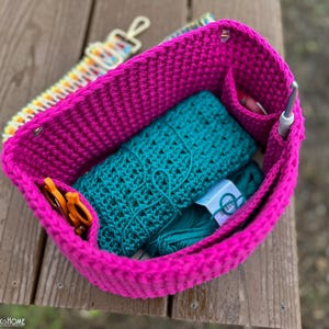 May include: A bright pink crochet project bag filled with teal yarn, a teal crochet wrap, orange scissors, and a crochet hook. A colorful strap and other notions are visible. The bag sits on a wooden surface.
