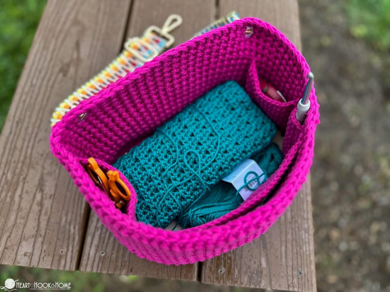 May include: A bright pink crochet project bag filled with teal yarn, a teal crochet wrap, orange scissors, and a crochet hook. A colorful strap and other notions are visible. The bag sits on a wooden surface.