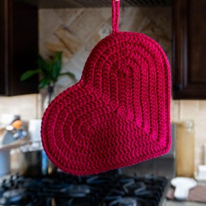 May include: A handmade, heart-shaped potholder in a vibrant shade of red. The potholder is crafted with a crochet technique, featuring concentric circular patterns. It has a loop for hanging and is designed for kitchen use.