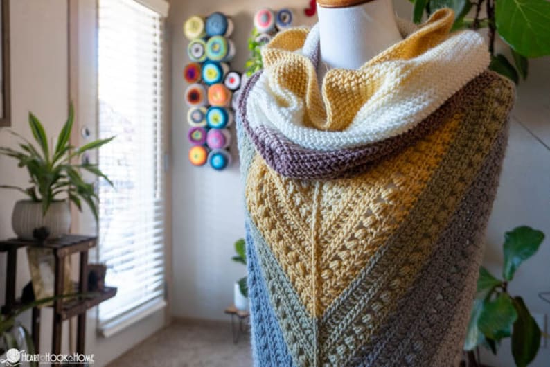 May include: A crocheted shawl with a multi-colored chevron pattern in shades of blue, green, yellow, and brown. The shawl is draped over a white mannequin and has a matching crocheted cowl.