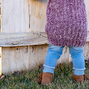 May include: A child wearing a pink fuzzy sweater and blue jeans stands in front of a weathered wooden fence. The child is wearing brown boots with fringe.