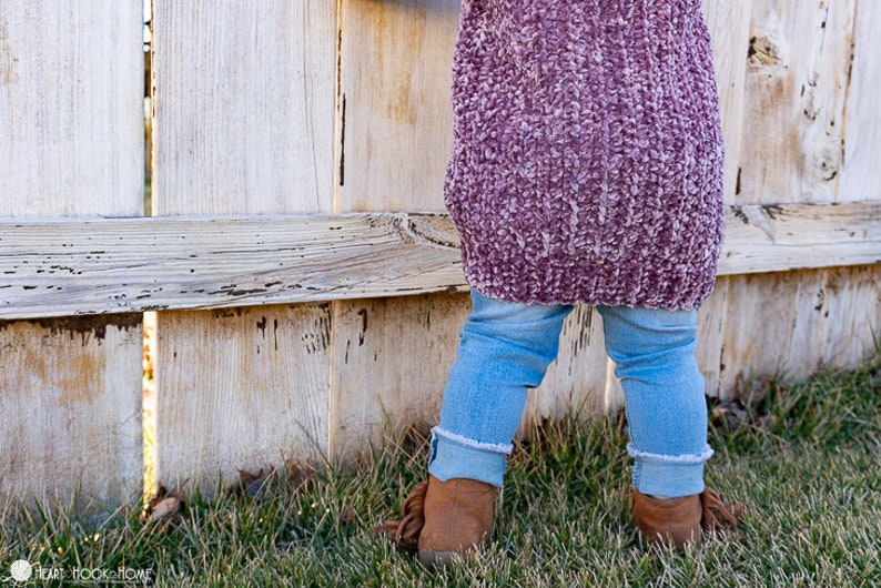 May include: A child wearing a pink fuzzy sweater and blue jeans stands in front of a weathered wooden fence. The child is wearing brown boots with fringe.