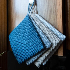 May include: Four crocheted pot holders hanging on a hook. The pot holders are in shades of blue and grey.
