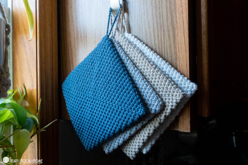 May include: Four crocheted pot holders hanging on a hook. The pot holders are in shades of blue and grey.