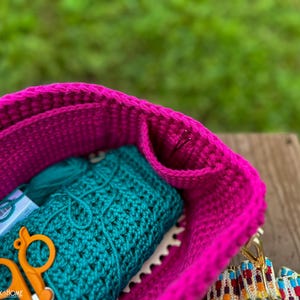May include: A close-up of a magenta crochet bag filled with teal yarn, a crocheted teal item, and orange scissors. The bag has a textured appearance and a colorful strap. The background is blurred green.